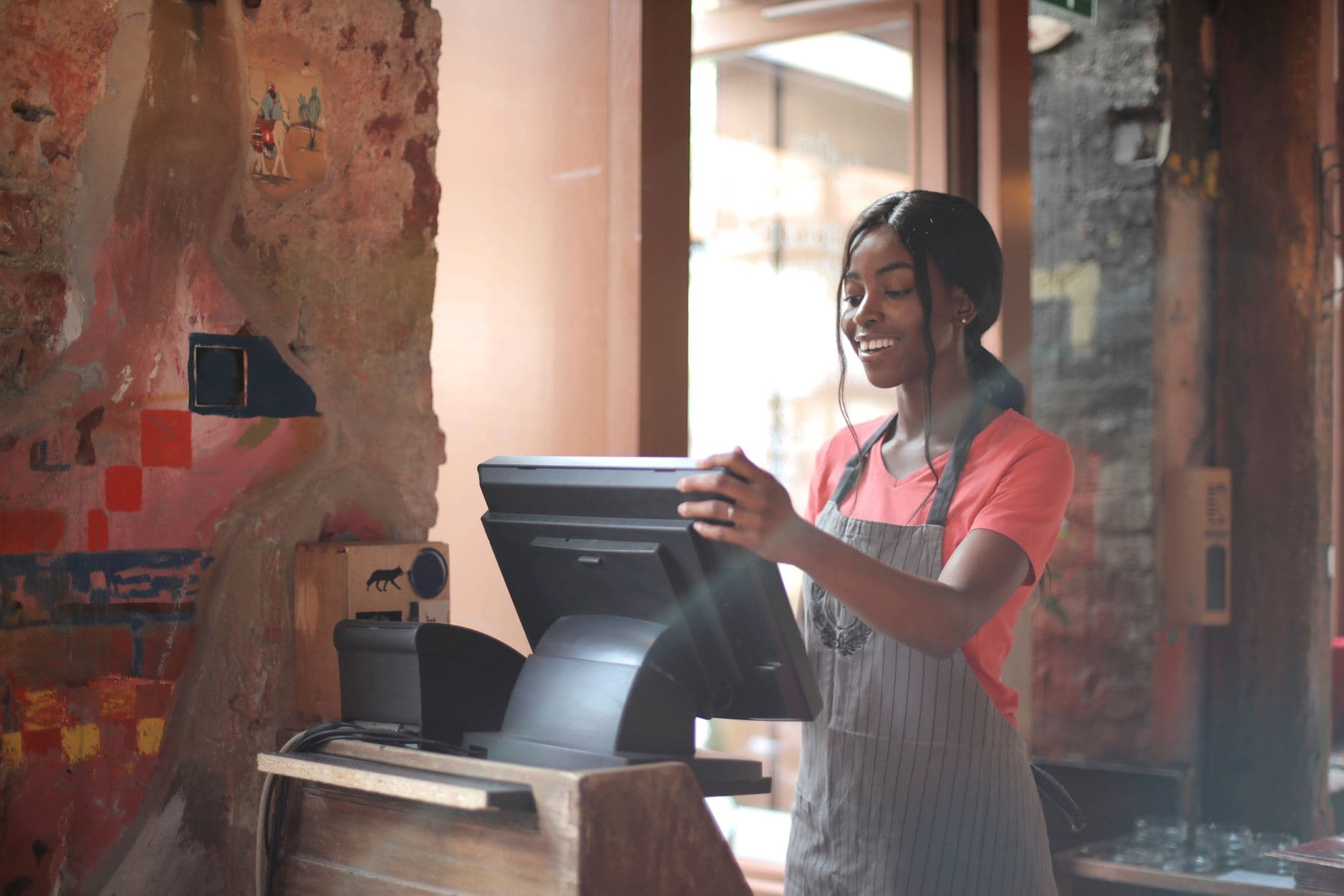 Cheerful American African waitress in apron working on counter monitor while registering order at cozy cafe.