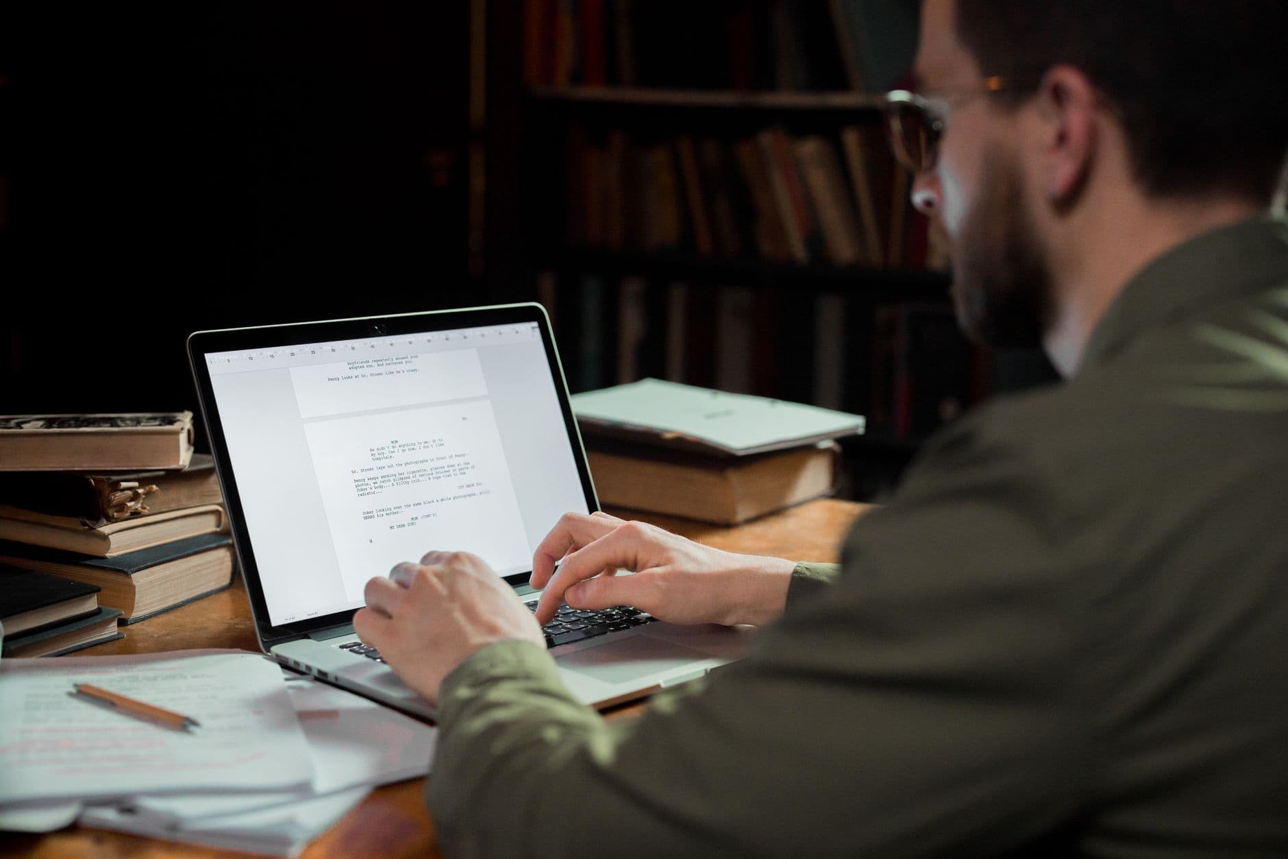 A man focuses on typing a document in a warm, cozy library environment, surrounded by books.