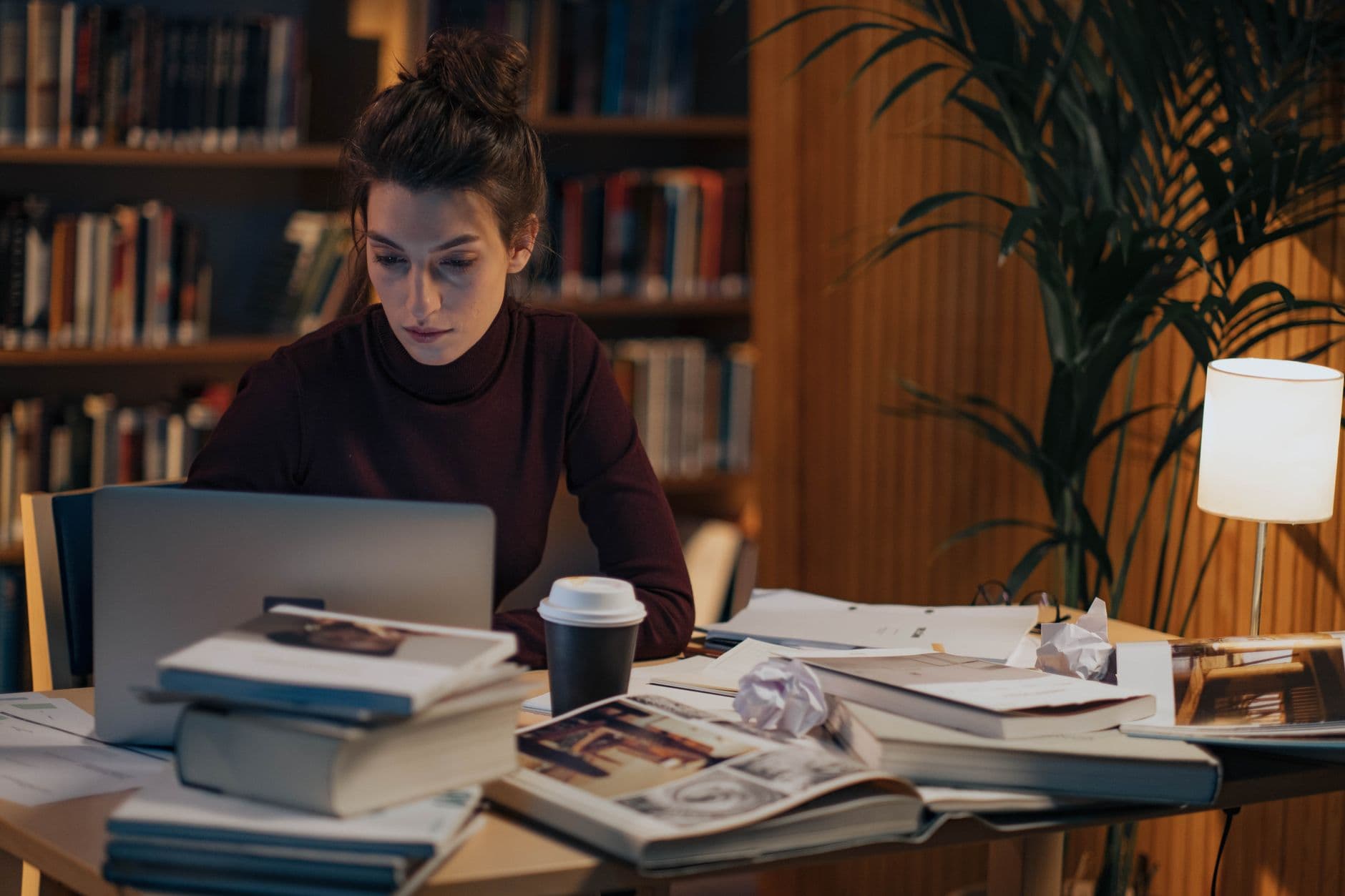 A woman working late at a desk covered in books, a laptop and coffee.